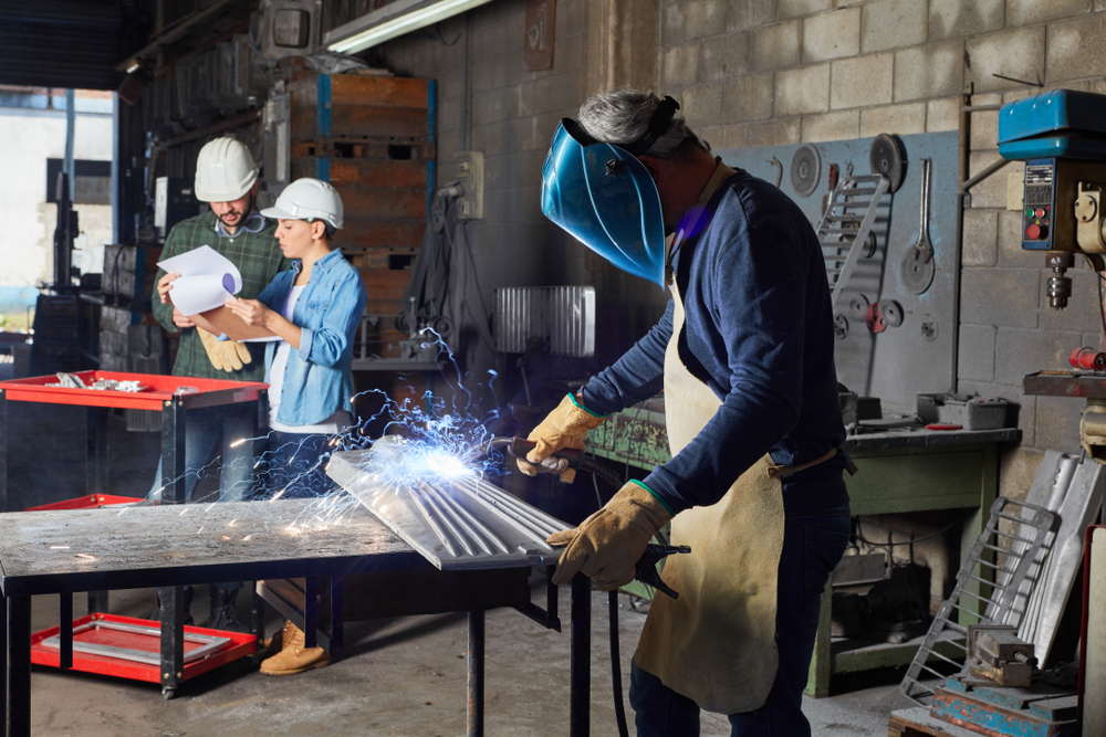 Team reviewing project plans while a welder works, showing capabilities to consider when comparing metal fabrication companies.